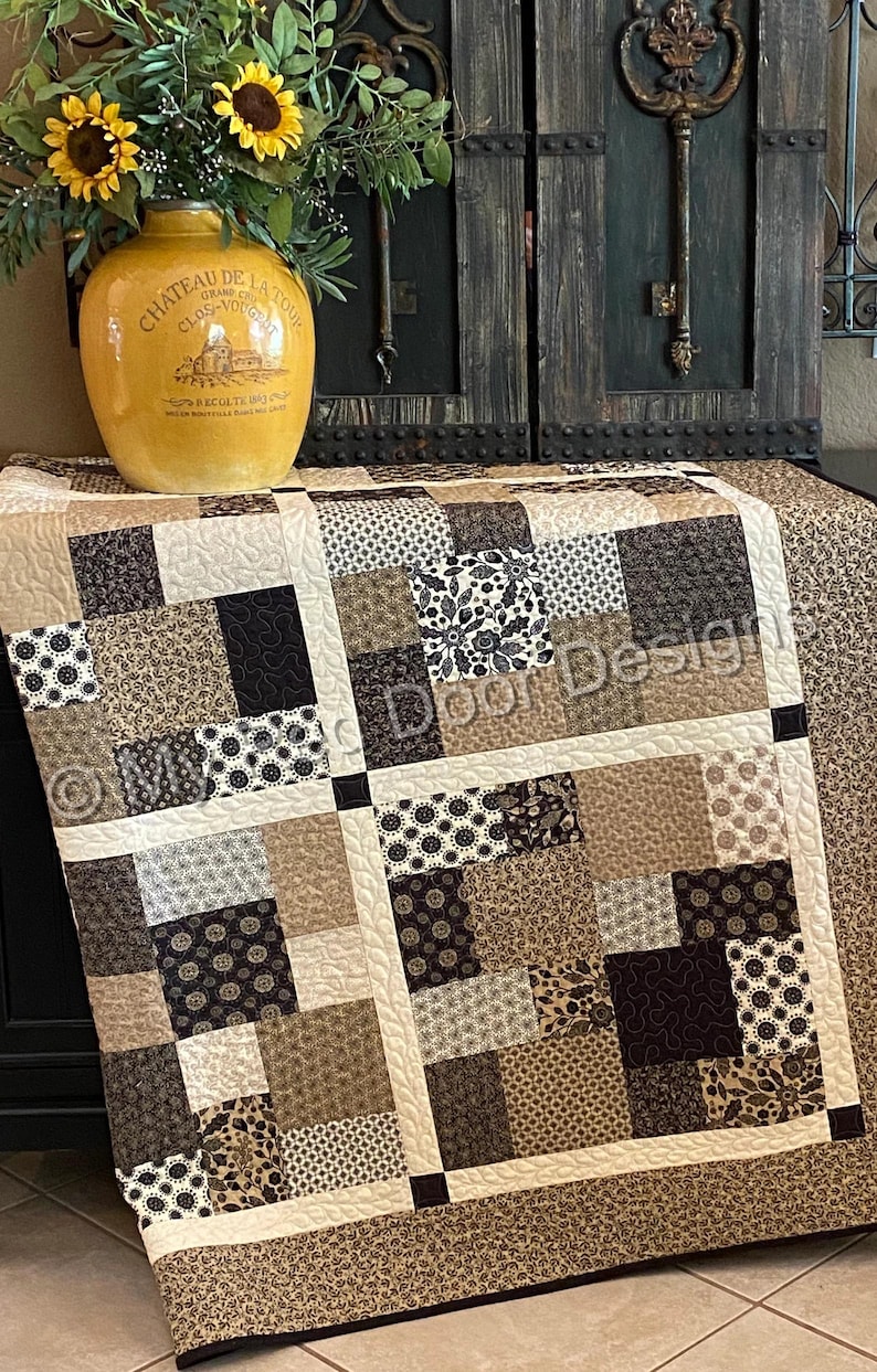 Op de afbeelding: Een patchwork quilt met een raster van vierkante stoffen blokken in tinten zwart, bruin, beige en cr&egrave;me. De quilt toont diverse patronen waaronder bloemmotieven, geometrische vormen en krulontwerpen, gescheiden door cr&egrave;mekleurige stroken.