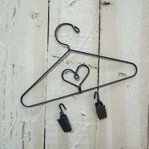 Może przedstawiać: Czarny metalowy wieszak druciany z motywem serca pośrodku. Dwa małe czarne klipsy wiszą poniżej wieszaka. Wieszak jest prezentowany na białym, zwietrzałym drewnianym tle.
