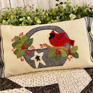 May include: A cream-coloured pillow with a black and white striped border. The pillow features an embroidered design of a red cardinal perched on a branch with green leaves and red berries. A black and white star is in a basket behind the cardinal.