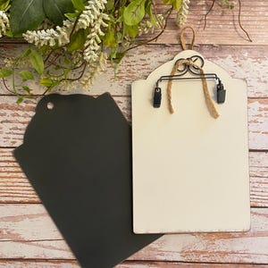 May include: Two rectangular wooden plaques with rounded tops. One plaque is painted matte black with a circular hole at the top. The other plaque is painted off-white, featuring a black metal clip mechanism attached with twine for holding items.