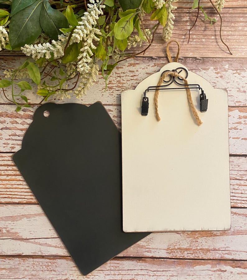 May include: Two rectangular wooden plaques with rounded tops. One plaque is painted matte black with a circular hole at the top. The other plaque is painted off-white, featuring a black metal clip mechanism attached with twine for holding items.