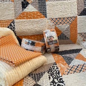 Puede incluir: Una colcha de retazos con patrones geom&eacute;tricos en naranja, negro, blanco y gris con tela crema texturizada. Dos rollos de tiras de tela est&aacute;n envueltos en pl&aacute;stico transparente, etiquetados con Moda Fabrics y un gr&aacute;fico de cinta m&eacute;trica.