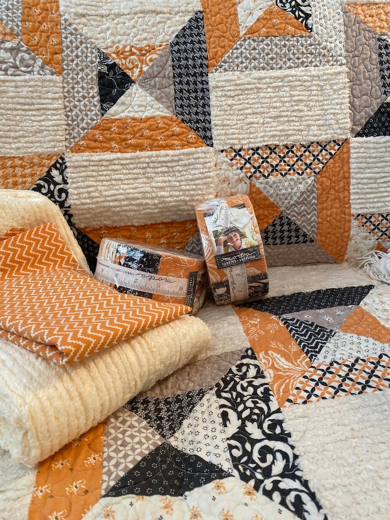 Puede incluir: Una colcha de retazos con patrones geom&eacute;tricos en naranja, negro, blanco y gris con tela crema texturizada. Dos rollos de tiras de tela est&aacute;n envueltos en pl&aacute;stico transparente, etiquetados con Moda Fabrics y un gr&aacute;fico de cinta m&eacute;trica.