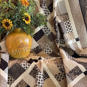 Op de afbeelding: Een patchwork quilt met diverse bruine, beige en cr&egrave;mekleurige stoffen vierkanten met bloem- en geometrische patronen, geaccentueerd door een gele keramische kan met zonnebloemen en de tekst My Red Door Designs op de voorzijde.