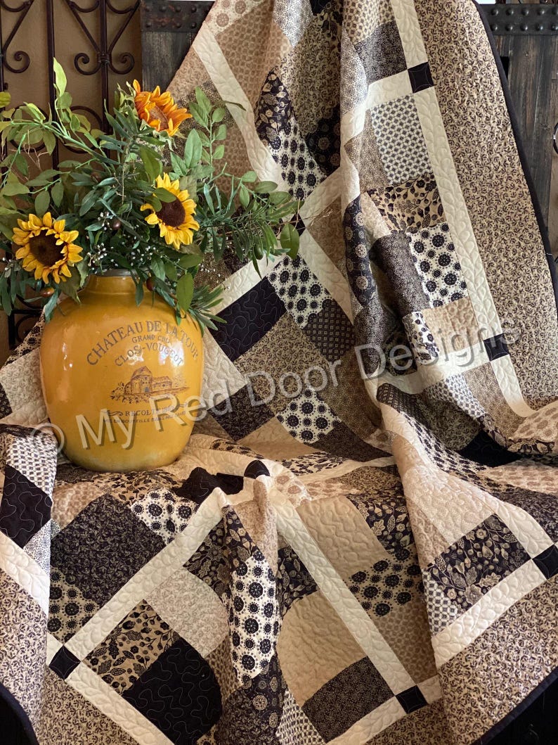 Op de afbeelding: Een patchwork quilt met diverse bruine, beige en cr&egrave;mekleurige stoffen vierkanten met bloem- en geometrische patronen, geaccentueerd door een gele keramische kan met zonnebloemen en de tekst My Red Door Designs op de voorzijde.