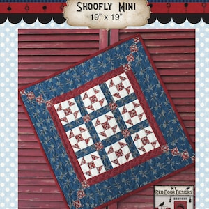 May include: A red, white, and blue quilt block with a "Shoofly Mini" title and the measurement 19" x 19". The quilt block is on a red wooden background. The quilt block is made with a blue fabric with white stars, a red fabric with white squares, and a white fabric with red squares. The quilt block is surrounded by a red border and a blue border. The quilt block is made by My Red Door Designs.