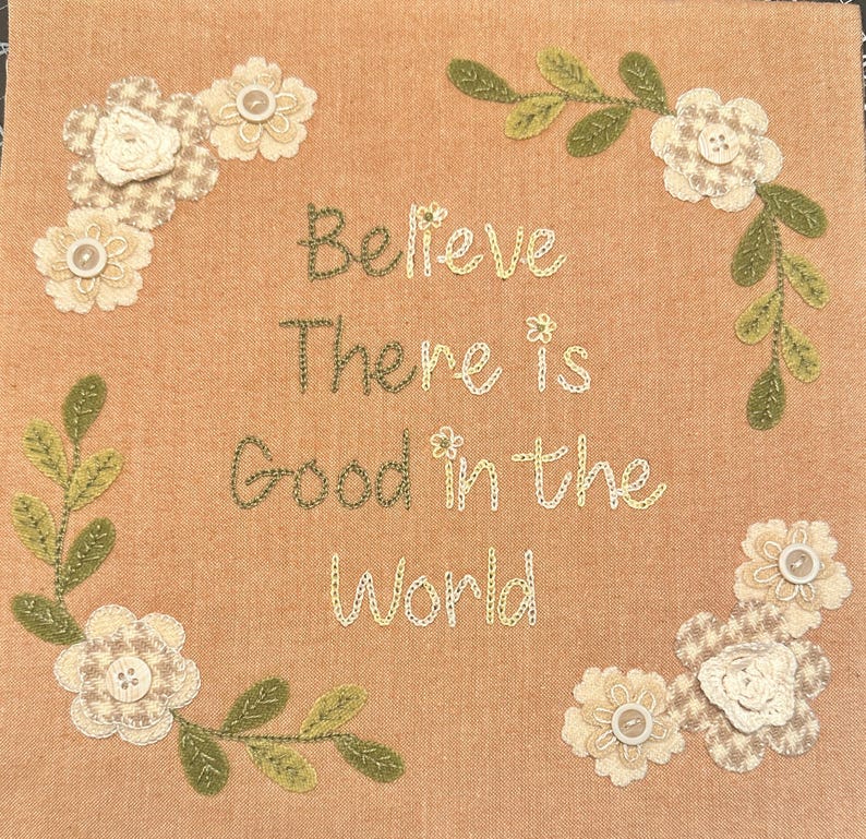 May include: Beige fabric with embroidered green vines and white flowers. The text "Believe there is good in the world" is embroidered in the center.