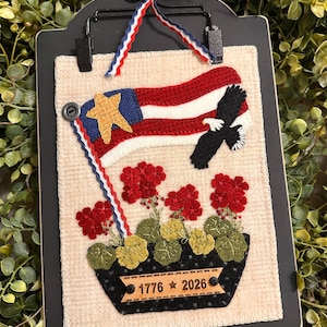 May include: A patriotic wall hanging featuring a stylized American flag with a gold star and a black eagle. Red flowers and green foliage are in a black planter with the dates 1776 and 2026. The piece is framed in black and has a red, white, and blue ribbon.