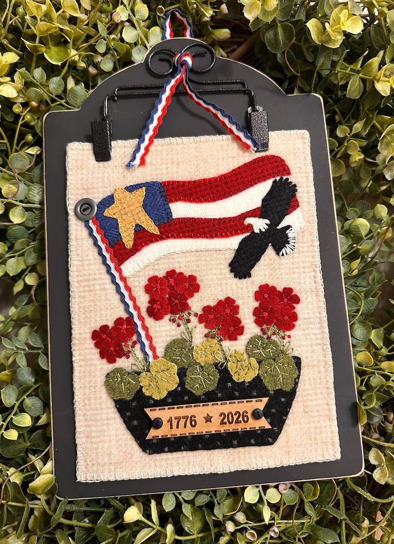 May include: A rectangular fabric wall hanging featuring a beige waffle-textured background. It displays an embroidered American flag, a black eagle, and red geraniums in a black planter. A wooden plaque reads 1776 2026 with a star.