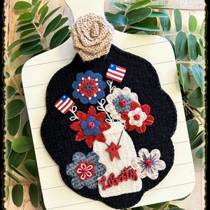 May include: A white wooden cutting board with a black felt background featuring a felt flower arrangement in a mason jar. The flowers are red, white, and blue with red buttons for centers. The mason jar has a red star and the word "Liberty" written on it. There are two small American flags in the arrangement.
