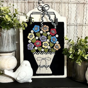 May include: A colorful felt flower bouquet in a white and black checkered vase with a white ribbon. The bouquet is displayed on a black background with a white frame.