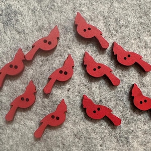 May include: A collection of eight red wooden bird-shaped buttons. Each button features a bird silhouette with two small holes for sewing. The buttons are scattered on a gray felt surface.