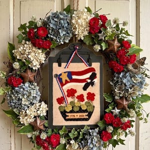 May include: A decorative wreath featuring blue, white, and red flowers, green leaves, and metal stars. A central black clipboard holds a rectangular fabric piece with a red, white, and blue flag, a black eagle, red flowers, and the text 1776 * 2026.