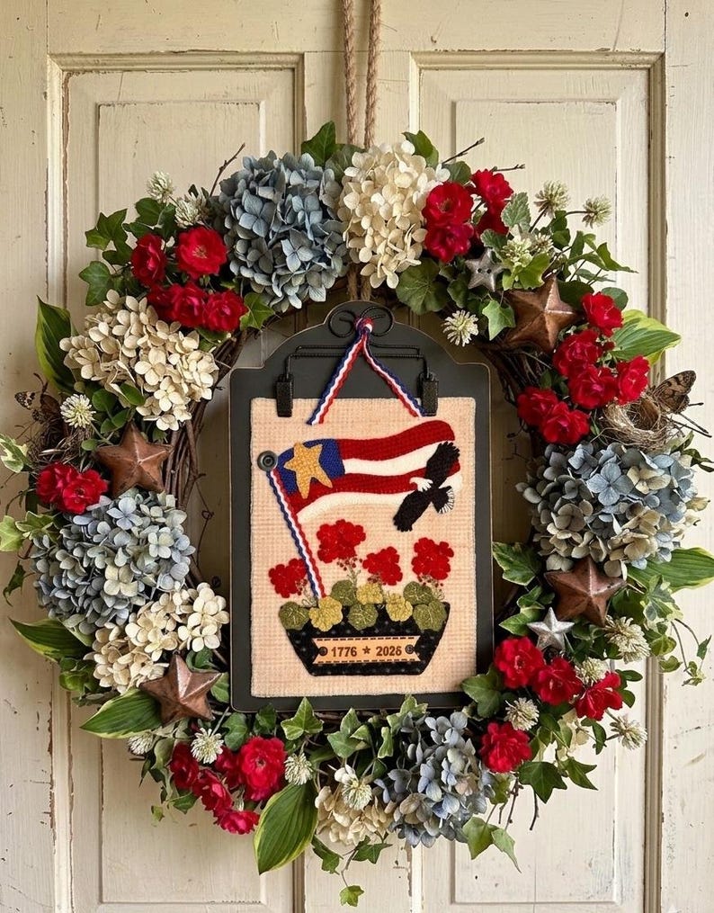 May include: A decorative wreath featuring blue, white, and red flowers, green leaves, and metal stars. A central black clipboard holds a rectangular fabric piece with a red, white, and blue flag, a black eagle, red flowers, and the text 1776 * 2026.