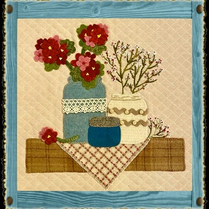 Könnte beinhalten: Ein gesteppter Wandbehang mit einer Stillleben-Darstellung von drei Vasen mit Blumen. Die Vasen sind mit Spitze, Knöpfen und einem Jutedeckel verziert. Die Blumen sind rosa und rot und in einer Vase angeordnet. Der Hintergrund ist hellrosa.