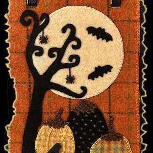 May include: A Halloween-themed felt wall hanging. The design features a full moon, bats, a silhouetted tree with spiders, and pumpkins. The background is orange with a plaid pattern. A black ribbon is attached for hanging.