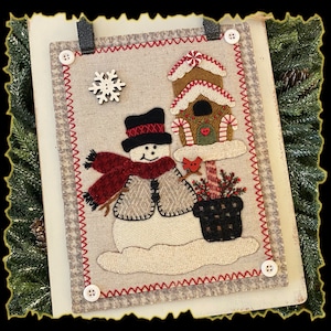 May include: A felt wall hanging featuring a snowman, a gingerbread house, and a potted plant. The snowman is wearing a red scarf and a black hat. The gingerbread house has candy canes and a red heart on the door. The potted plant has red berries and a red bird.
