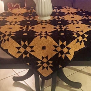 Puede incluir: Un camino de mesa acolchado negro y dorado con un patrón de estrellas. El camino de mesa está sobre una mesa redonda con dos sillas negras.