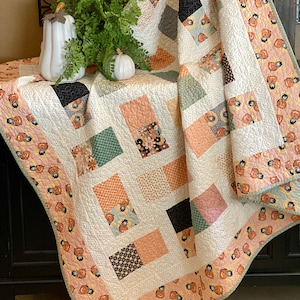 May include: A white quilt with a patchwork design featuring squares of various floral and geometric patterns in shades of orange, green, black, and white. The quilt is folded and draped over a dark brown wooden surface.