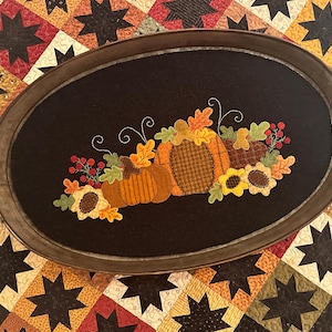 Pode incluir: Uma bandeja oval preta com uma borda de metal marrom, apresentando um design bordado de abóboras, girassóis e folhas em tons de laranja, amarelo e verde. A bandeja é perfeita para servir lanches ou bebidas.