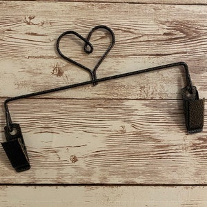 Può includere: Un portasciugamani in metallo nero a forma di cuore con due clip su ogni estremità. Il portasciugamani è montato su una parete di legno bianca.