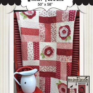 May include: A red, white, and pink quilt with a floral design. The quilt is 50 inches by 58 inches and is called "Whimsy Flowers". The quilt is displayed on a red wooden shutter with a white pitcher and a red chair in the foreground.