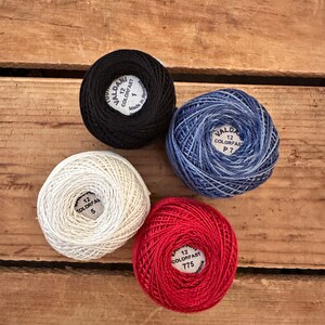 May include: Four spools of embroidery floss in black, white, red, and blue. Each spool has a label with the brand name "Valdani" and the text "12 Colorfast" followed by a number.