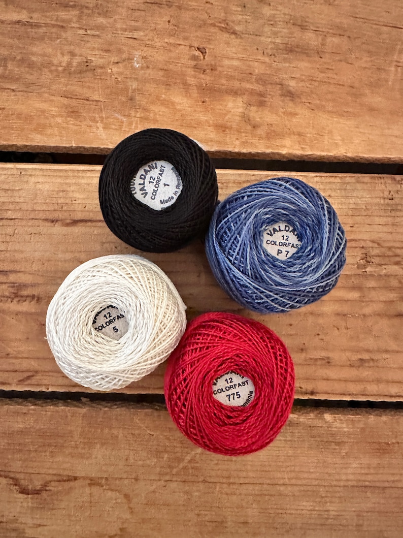 May include: Four spools of embroidery floss in black, white, red, and blue. Each spool has a label with the brand name "Valdani" and the text "12 Colorfast" followed by a number.