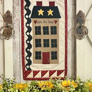 May include: A quilted wall hanging featuring a house design. The house is beige with black windows and a red door. The roof is black with two yellow stars. The text "Bless this House" is embroidered on the front of the house. The hanging is surrounded by a red and white border.