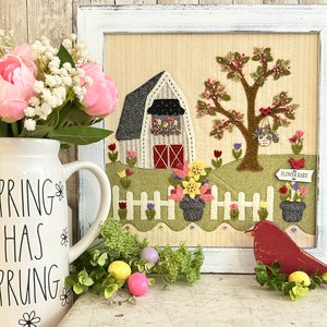 May include: A white pitcher with the text "Spring Has Sprung" and black floral accents, a framed needlepoint design of a red barn with a white picket fence, a flowering tree, and a red bird, and a red wooden bird figurine with a nest of green leaves and yellow eggs.