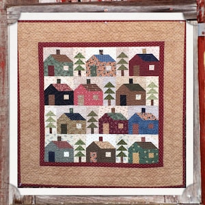 May include: A square quilt titled Pine Ridge 33" Square by My Red Door Designs. The quilt features a grid pattern of various houses and pine trees in assorted colors and fabric prints, surrounded by a dark red border and a tan outer border.
