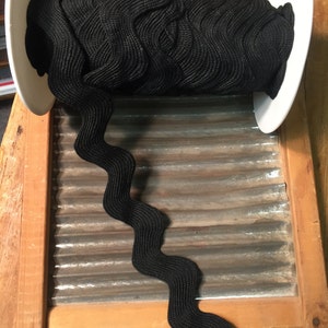 May include: A spool of black, wavy, elastic trim. The trim is wound around a white cardboard spool with the text "National Washboard Co. No. National 442 Made in U.S.A." printed on it. The spool is sitting on a wooden washboard.