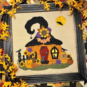 May include: A felt wall hanging with a witch's hat shaped house. The house has a red door, a window with two cats inside, and a sign that reads "The Critter Corner". The house is surrounded by sunflowers, pumpkins, and a black cat.