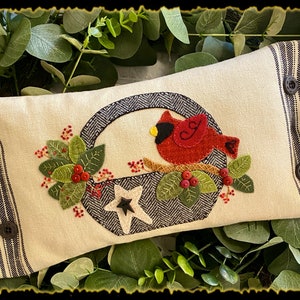 May include: A white pillow with a black and white checkered border. The pillow features an embroidered design of a red cardinal perched on a branch inside a basket with a white star. The design also includes green leaves and red berries.