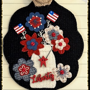 May include: A felt wall hanging with a black background featuring a white mason jar with red stars and the word "Liberty" in red. The jar is filled with red, white, and blue flowers and small American flags.
