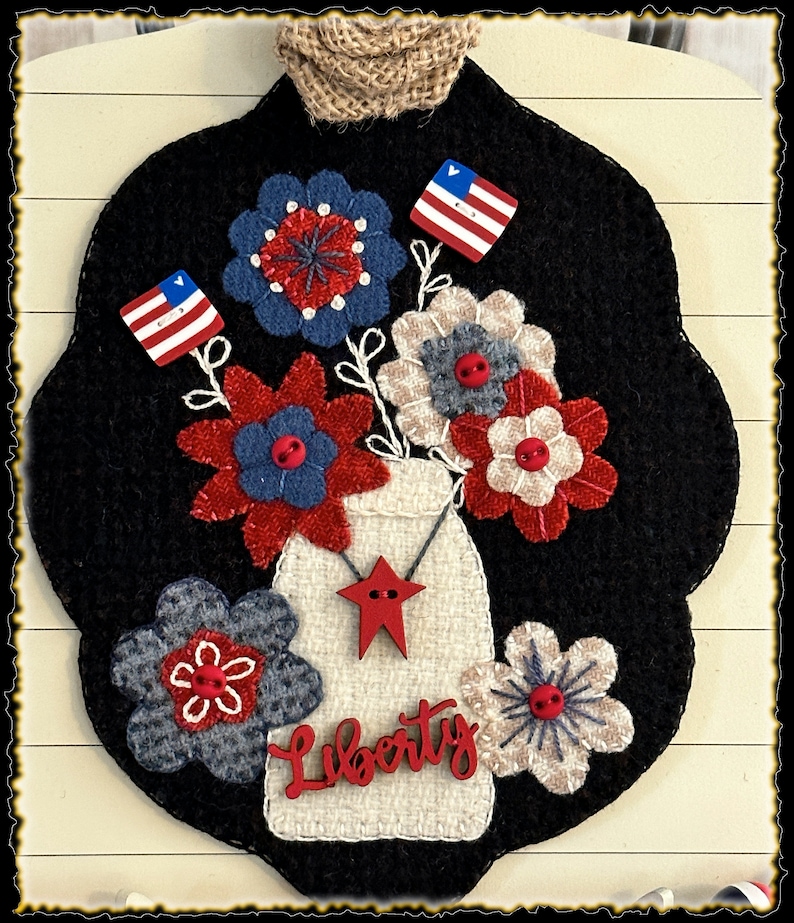 May include: A felt wall hanging with a black background featuring a white mason jar with red stars and the word "Liberty" in red. The jar is filled with red, white, and blue flowers and small American flags.