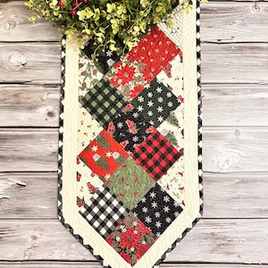 Könnte beinhalten: Ein gesteppter Tischläufer mit Weihnachtsmotiv. Der Läufer besteht aus verschiedenen Stoffen mit roten, grünen, schwarzen und weißen Mustern. Der Läufer hat einen weißen Rand mit einem schwarz-weiß karierten Besatz.