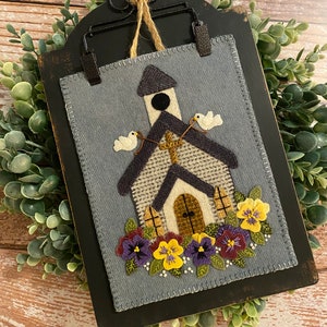 May include: A felt wall hanging with a blue background featuring a white church with a cross, two white doves, and purple and yellow flowers. The church is framed in a black wooden frame.