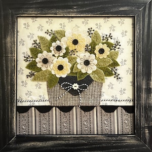 May include: Framed textile art featuring a floral arrangement. The bouquet includes cream and yellow flowers with black button centres, green leaves, and a grey basket. The background is cream fabric with small floral patterns. The frame is black.