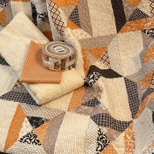 Puede incluir: Una colcha de patchwork con un patr&oacute;n geom&eacute;trico en tonos naranja, crema, marr&oacute;n y negro. La colcha est&aacute; superpuesta con una pieza doblada de tela de chevr&oacute;n naranja y un rollo redondo de tiras de tela atado con una banda transparente.