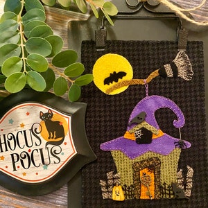 May include: A black felt Halloween door hanger featuring a witch's house, a broom, a moon, and bats. The house has a purple hat, a green door, and a key. The text "Hocus Pocus" is on a separate black felt sign.