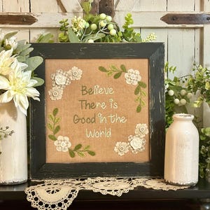 May include: A framed embroidery with a burlap background and white embroidery thread. The embroidery features a wreath of green leaves and white flowers with the text "Believe There is Good in the World". The frame is black with a distressed finish. The frame is sitting on a shelf with two white ceramic vases and green plants.