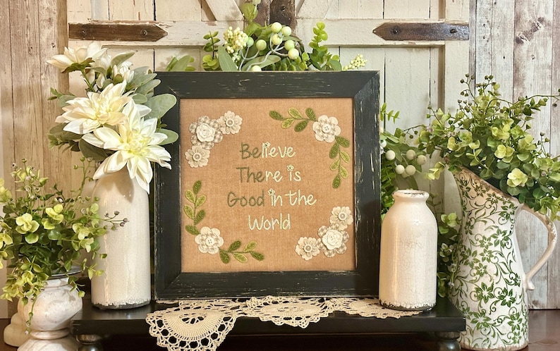 May include: A framed embroidery with a burlap background and white embroidery thread. The embroidery features a wreath of green leaves and white flowers with the text "Believe There is Good in the World". The frame is black with a distressed finish. The frame is sitting on a shelf with two white ceramic vases and green plants.