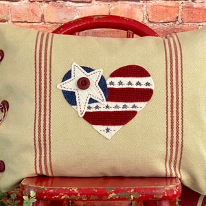 May include: A beige pillow with red stripes and a red, white, and blue heart-shaped applique with a star. The pillow has red buttons and ties.