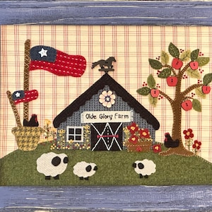 May include: A framed folk art piece with a blue wooden frame. The artwork depicts a barn with the text "Olde Glory Farm", a tree with red apples, sheep, and American flags. The background is a plaid pattern.