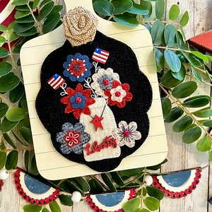 May include: A felt craft depicting a mason jar filled with red, white, and blue flowers. The jar is labeled "Liberty" and has a red star on the side. The craft is mounted on a wooden cutting board with a burlap bow.