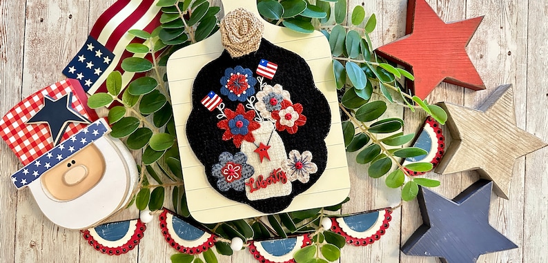 May include: A felt craft depicting a mason jar filled with red, white, and blue flowers. The jar is labeled "Liberty" and has a red star on the side. The craft is mounted on a wooden cutting board with a burlap bow.