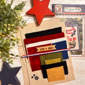 May include: A craft kit featuring a tan burlap background, assorted fabric swatches in black, red, white, blue, olive, and mustard, a red wooden star, a blue wooden star, and a wooden tag printed with 1776 and 2026, plus a pattern instruction sheet.