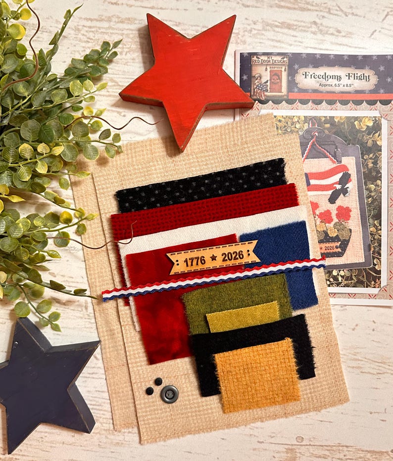 May include: A craft kit featuring a tan burlap background, assorted fabric swatches in black, red, white, blue, olive, and mustard, a red wooden star, a blue wooden star, and a wooden tag printed with 1776 and 2026, plus a pattern instruction sheet.
