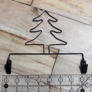 May include: Black metal Christmas tree-shaped clip with two attached clips. The tree is made of wire and is approximately 4 inches wide. The clips are black and are attached to a horizontal bar.